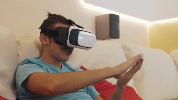 Young man sitting on sofa wears virtual reality headset makes money tossing gesture - Кадры, видео