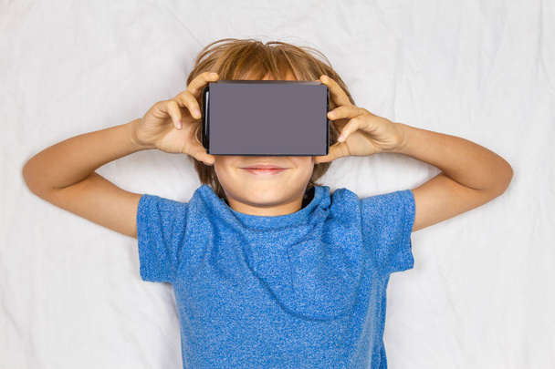Ребенок лежит в белой кровати с 3D виртуальной реальности, картонные очки VR
 - Фото, изображение