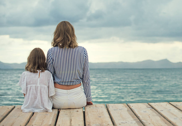 Woman and little girl sitting on the wooden pier near the sea. - Φωτογραφία, εικόνα