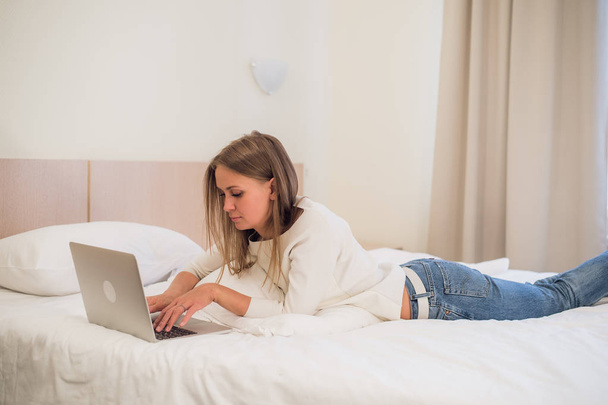 Mujer rubia atractiva pensativa usando una computadora portátil mientras se sienta en el borde de una cama en casa o en un dormitorio de hotel en vacaciones o viaje de negocios
 - Foto, Imagen