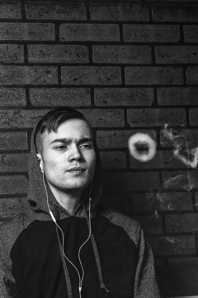 Young handsome white guy is admiting puffs of steam from the electronic cigarette. Vaping. Teenager listening to music. Black and white photo. - 写真・画像