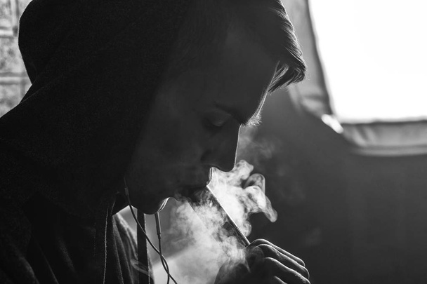 Young handsome white guy is admiting puffs of steam from the electronic cigarette. Vaping. Teenager listening to music. Black and white photo. - Photo, image