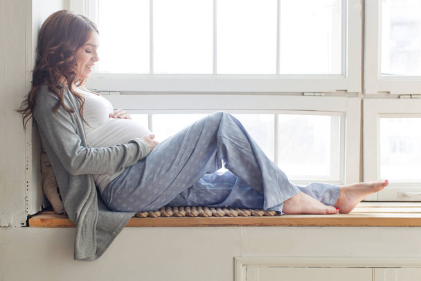 Mujer embarazada está sentada en el alféizar de la ventana
 - Foto, imagen