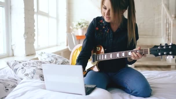 Attractive young girl learning to play electric guitar with notebook sit on bed in bedroom at home - Materiaali, video