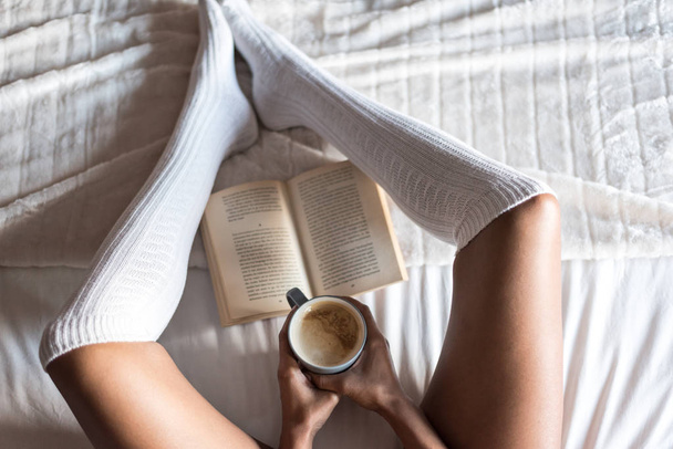 woman reading a book and drinking coffee on bed with socks - Photo, image