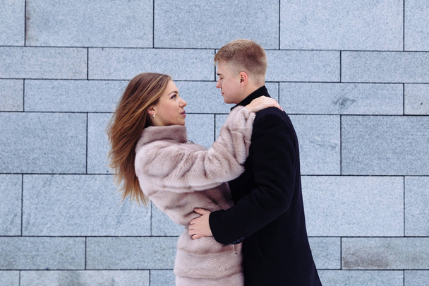 A young heterosexual man and woman in fur coat couple hug and kiss each other happy in a city. - Photo, Image