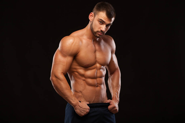 Strong Athletic Man - Fitness Model showing Torso with six pack abs. stands straight and puts his hands in trousers. isolated on black background with copyspace. - Photo, image