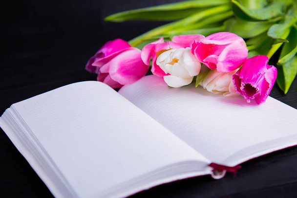 Bouquet of tender pink tulips and open notebook on black wooden  - Foto, Imagen