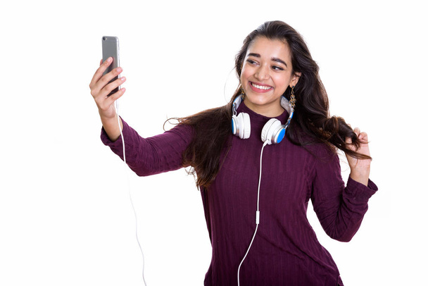Joven mujer india feliz sonriendo mientras usa auriculares alrededor del cuello y tomando foto selfie con teléfono móvil - Foto, Imagen