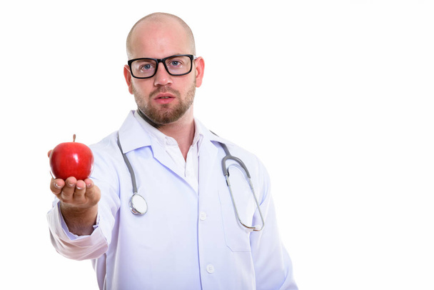 Studio shot of young bald muscular man doctor giving red apple - Zdjęcie, obraz
