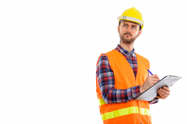 Studio shot of young man construction worker writing on clipboard while thinking - Photo, Image