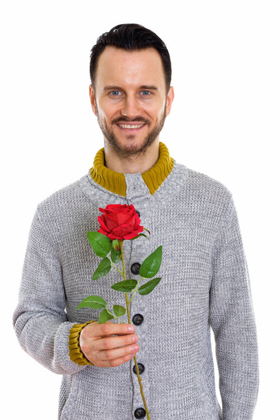 Studioaufnahme eines glücklichen jungen Mannes, der lächelt, während er eine rote Rose für den Valentinstag bereit hält - Foto, Bild