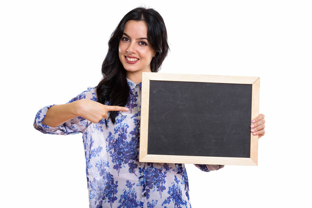 Studio shot of young happy Spanish businesswoman smiling while holding and pointing at blank blackboard - Φωτογραφία, εικόνα