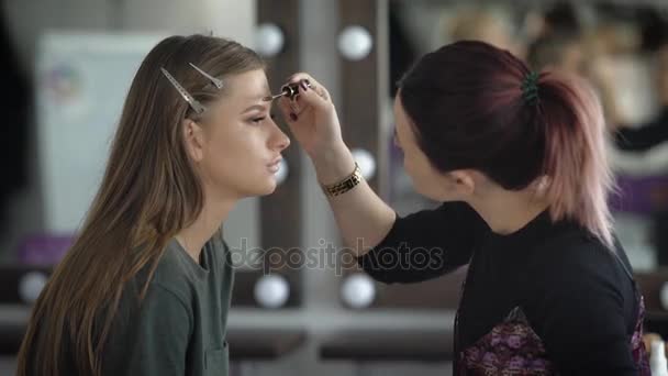 Makeup artist emphasizes the shape of eyebrows model using brown eyebrow gel. Model is sitting quietly. Her face has already been done moisturizer, foundation, concealer, eye liner, black mascara - Πλάνα, βίντεο
