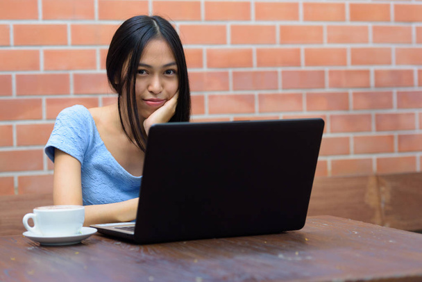 junge schöne asiatische Teenager Mädchen ruht Kinn auf der Hand mit Laptop und Cappuccino auf Holztisch gegen Ziegelwand - Foto, Bild