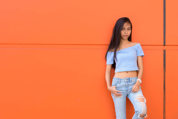 Giovane bella ragazza asiatica adolescente posa e appoggiata contro la parete dipinta arancione
 - Foto, immagini
