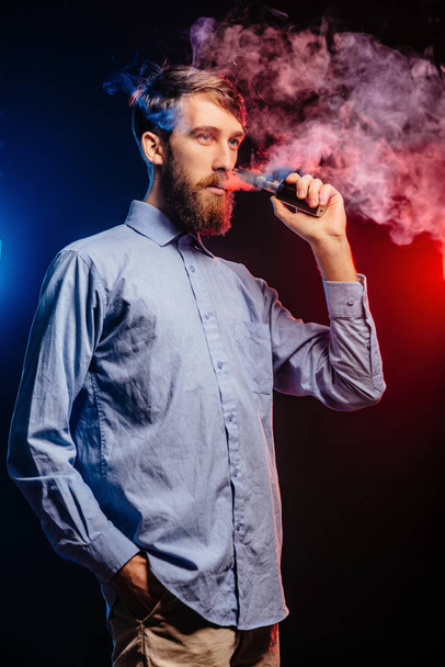 Young guy with a beard vaping and releases a cloud of vapor. hipster vaper smoke vaporizer. - Photo, Image
