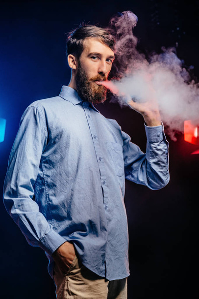 Young guy with a beard vaping and releases a cloud of vapor. hipster vaper smoke vaporizer. - Photo, Image