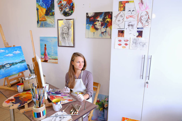 Κορίτσι που κάθεται σε σκαμνί στο καβαλέτο και ζωγραφική γράφοντας, χρησιμοποιεί τη βούρτσα για να - Φωτογραφία, εικόνα