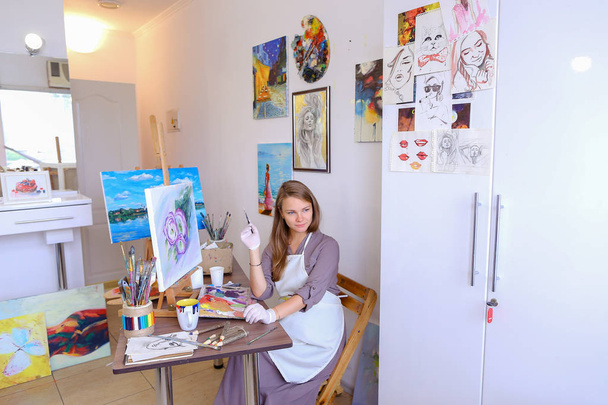 Κορίτσι που κάθεται σε σκαμνί στο καβαλέτο και ζωγραφική γράφοντας, χρησιμοποιεί τη βούρτσα για να - Φωτογραφία, εικόνα