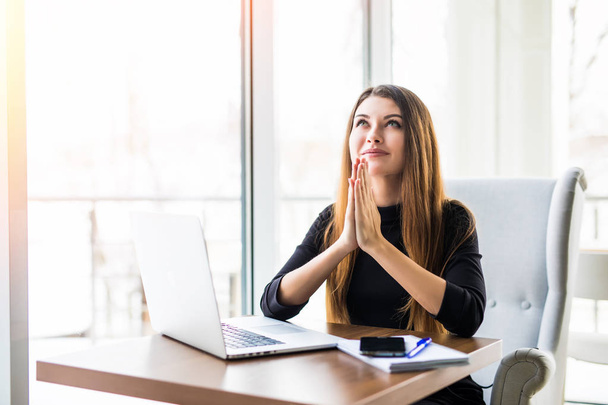 Geschäftsfrau betet. nachdenkliche Frau in Formalbekleidung, die die Hände umklammert hält und neben dem Laptop aufblickt, während sie an ihrem Arbeitsplatz sitzt - Foto, Bild
