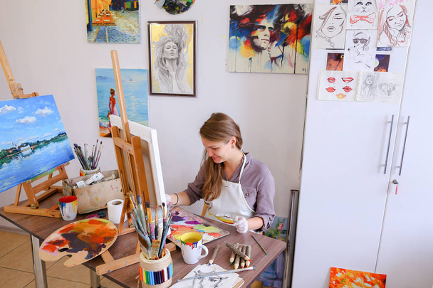 Chica se sienta en el taburete en el caballete y escribir pintura, utiliza el pincel para
 - Foto, Imagen