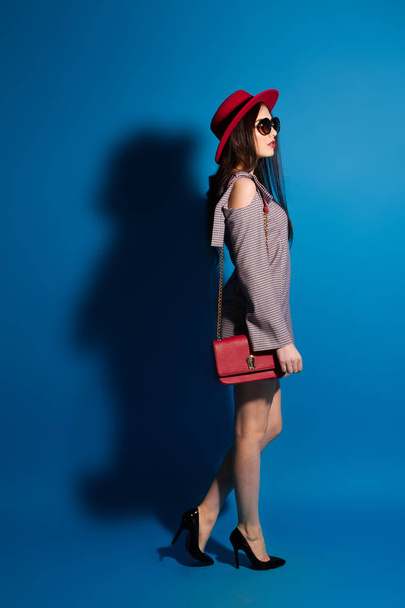 Lovely model in fashionable red Hat and a red Clutch on blue background. - Valokuva, kuva