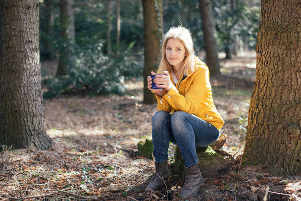 Милая блондинка-путешественница отдыхает в лесу и пьет горячий кофе или чай из винтажной кружки
 - Фото, изображение