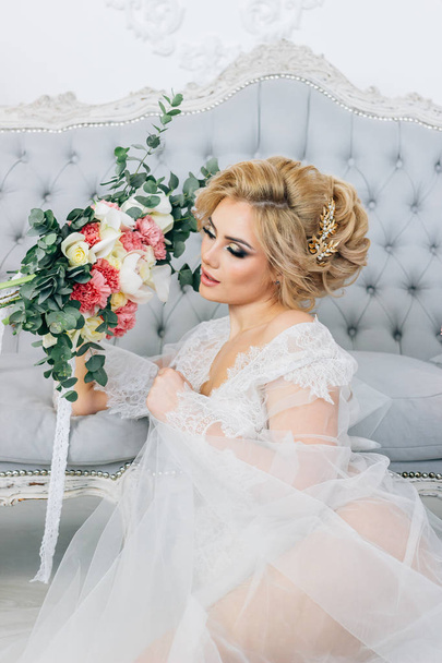 Portrait of a girl with a beautiful smile, make-up and hairstyle wearing a robe sitting on the floor holding a wedding bouquet - Fotografie, Obrázek
