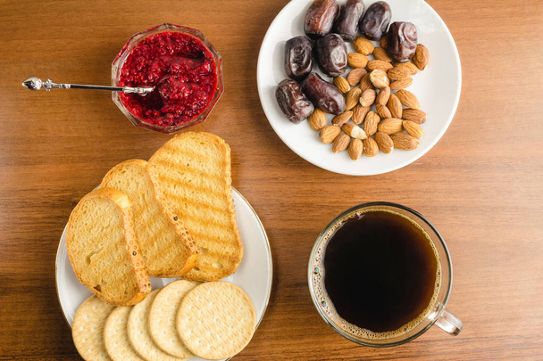 Breakfast time, cup of coffee with toasts, raspberry jam, dates, almonds on wooden table, top view - Photo, Image