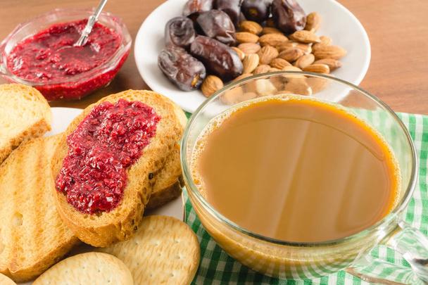 Frühstückszeit, Tasse Kaffee mit Milch, Toast, Himbeermarmelade, Datteln, Mandeln auf Serviette auf Holztisch - Foto, Bild