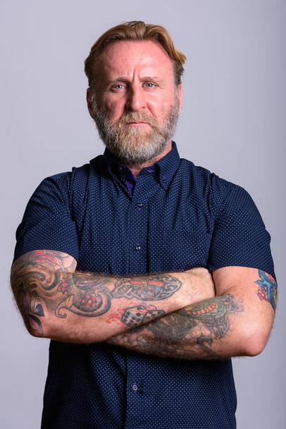 Зрелый бородатый мужчина с татуировками на руках, скрещенными в сером.
 - Фото, изображение