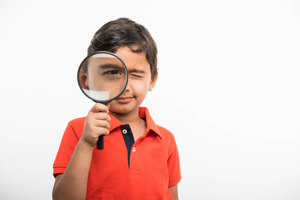 indisches Schulkind, das ein großes Auge und ein geschlossenes Auge zeigt und durch ein Vergrößerungsglas schaut - Foto, Bild