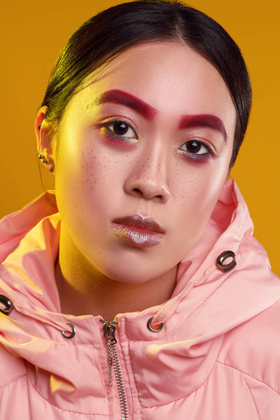 Portrait of an Asian girl with red eyebrows and make-up on a yellow background in the pink jacket with yellow light - Valokuva, kuva