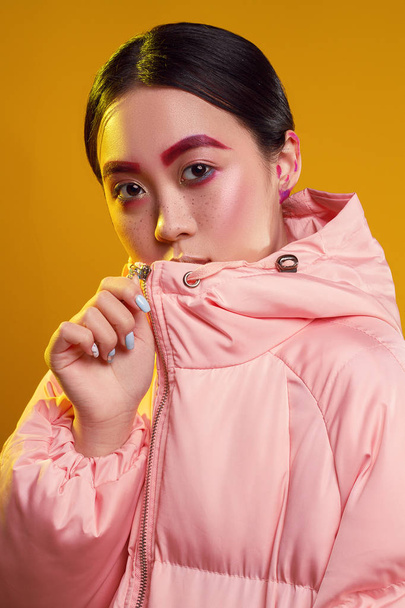 Retrato de una chica asiática con cejas rojas y maquillaje sobre un fondo amarillo en la chaqueta rosa con luz amarilla
 - Foto, Imagen
