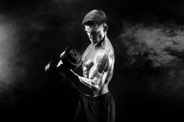 Handsome bodybuilder doing exercise with dumbbell. Studio shot. Black and white photo. Smoke - Foto, Imagen