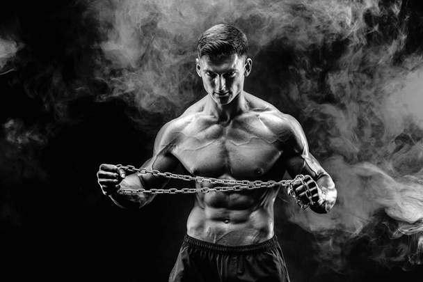 Portrait of muscular sportsman tearing metal chain.Black background with smoke - Фото, зображення