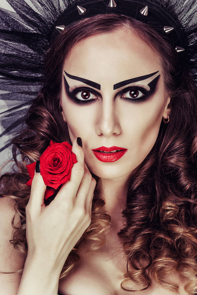 όμορφη γυναίκα με δραματικό μακιγιάζ και κόκκινα χείλη. Κόκκινο λουλούδι τριαντάφυλλο. Κορίτσι με διακόσμηση στα μαλλιά της. Νεαρή κοπέλα κοιτάζοντας την κάμερα - Φωτογραφία, εικόνα