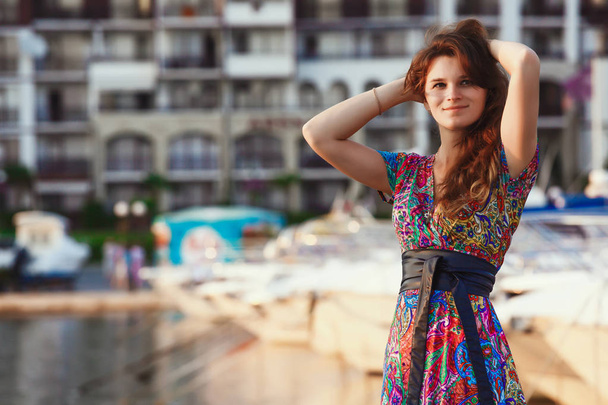 attractive brunette in long colorful dress standing alone on the beach against the backdrop of yachts and town at sunset - Fotó, kép