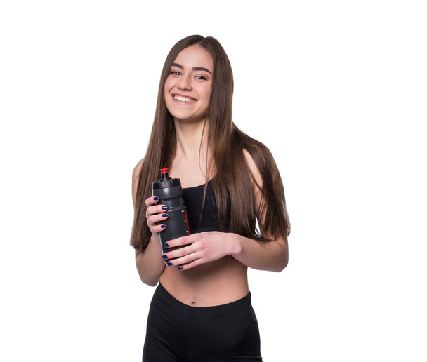 Portrait of smiling young sporty woman with a bottle of water posing in studio isolated on white background. - Фото, изображение