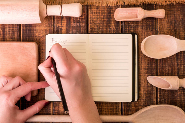 Vrouw hand schrijven een recept in het kookboek. Boek voor recept rond gebruiksvoorwerpen op houten achtergrond.  - Foto, afbeelding