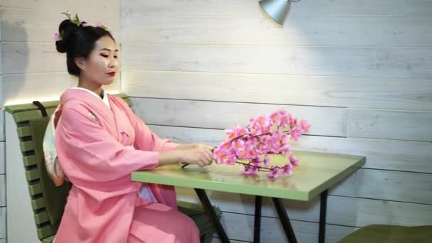 Prachtige geisha in traditionele Japanse kimono roze zitten aan een lege tafel in het restaurant en pakt op de takken van de kersenbloesem. - Video