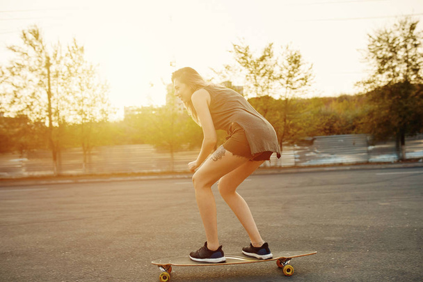 Beautiful young girl with tattoos riding longboard in sunny weather - Photo, Image