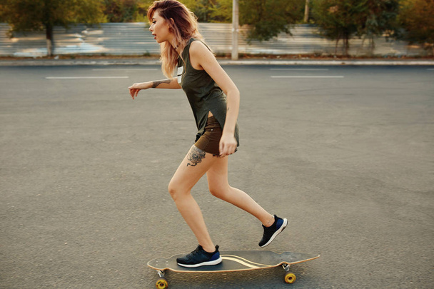 Menina bonita com tatuagens montando longboard em tempo ensolarado
 - Foto, Imagem