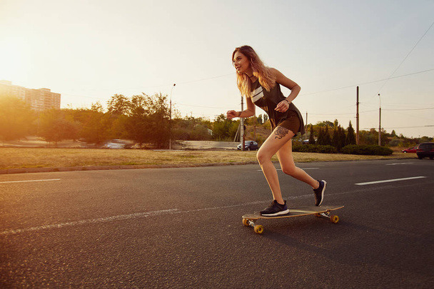 Beautiful young girl with tattoos riding longboard in sunny weather - Photo, Image
