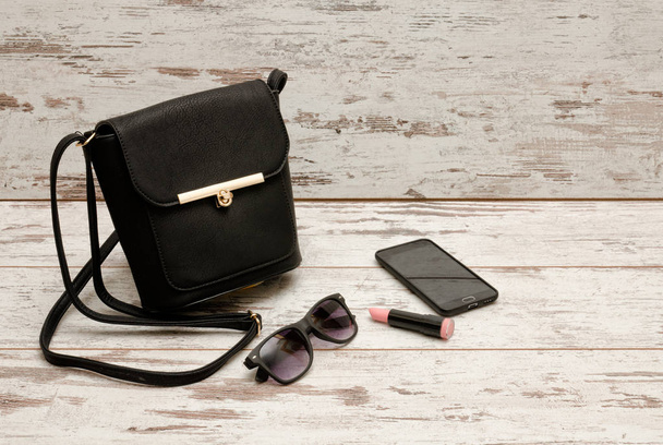 Bolso de mano para damas negras, gafas de sol, teléfono y lápiz labial sobre fondo de madera. concepto de moda
 - Foto, Imagen