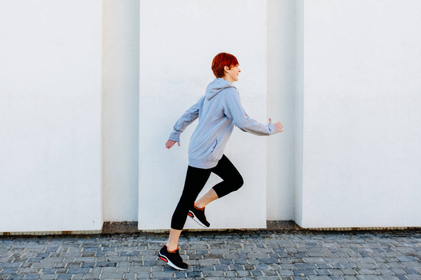 Jonge rode hoofd sport vrouw loopt langs de witte muur. - Foto, afbeelding