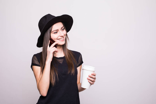 Portrét mladé dívky úsměv v klobouku držení mobilního telefonu a šálek kávy, samostatný na šedé - Fotografie, Obrázek