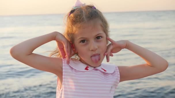 mooi klein meisje grimaces en tong toont op de achtergrond van de zonsondergang zee - Video
