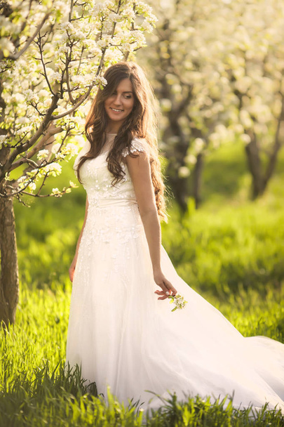 Portrait of beautiful woman in flowers. The bride in ivory wedding dress with long curly hair walking in gardens with summer blossom trees - Photo, image